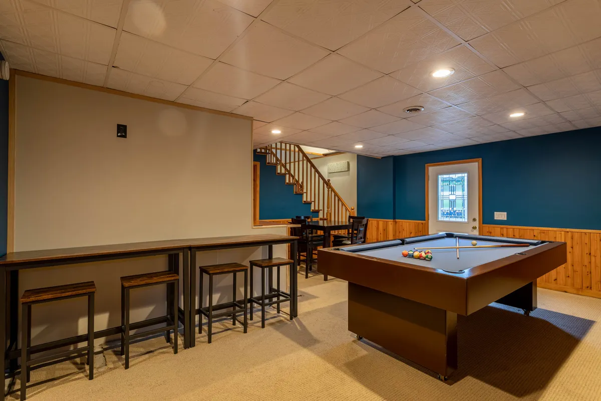 A vibrant interior scene centered on a pool table, bar seating, and a staircase. The room is set up for games, conversation, and casual dining—a lively hub for families to unwind together.