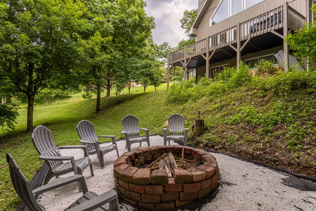 A cozy stone fire pit encircled by comfortable chairs is nestled among vibrant greenery. The area is set up for gatherings around the fire, with ample space for socializing, roasting marshmallows, or sharing stories under the stars.