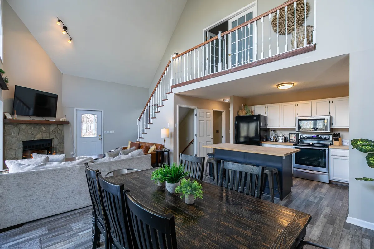 A spacious open-plan area featuring a central dining table, a sofa seating area with plush furnishings, a kitchen island visible in the background, a cozy fireplace, and a staircase. Warm lighting and wood accents give the space an inviting, communal atmosphere—perfect for large groups and family gatherings.