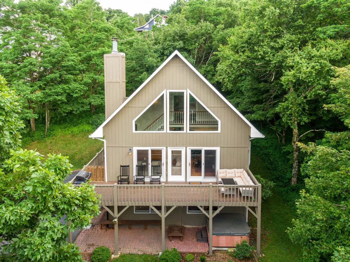 This hero image features the exterior of a chalet-style single-family home surrounded by lush greenery. Large windows span the front, flooding the interior with natural light, while an expansive deck invites guests to relax, dine, and gather outdoors. The elevated setting offers sweeping sunset views, perfect for stargazing or sipping morning coffee immersed in nature.