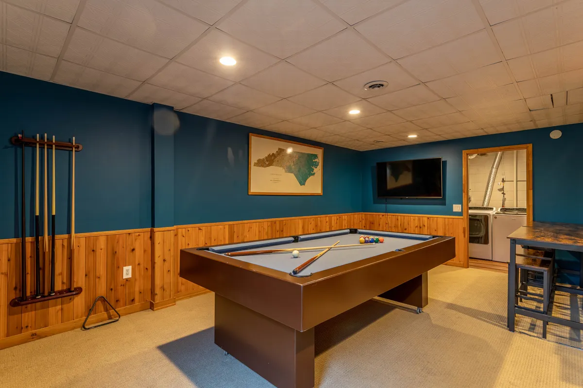 A vibrant game room featuring a full-size pool table as the central focus, a large flat screen TV mounted on the wall, stylish wall art, and a bar area in the background. The space is well-lit and designed for fun social gatherings and competitive play.