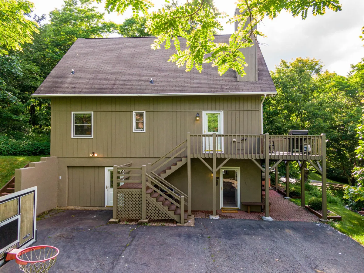 An exterior shot highlighting a spacious wooden deck with stairs, a basketball hoop, and large windows. The scene is framed by natural greenery, suggesting ample space for outdoor gatherings and activities.