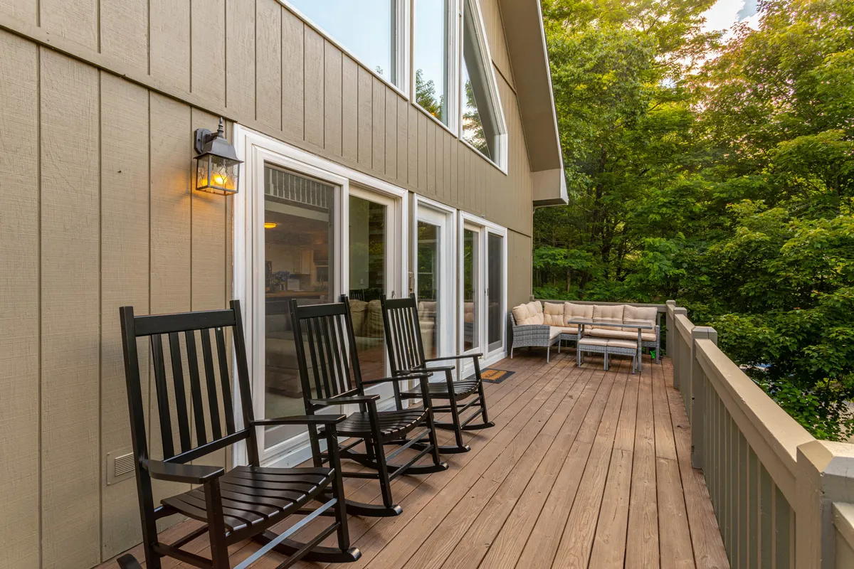 This image features a spacious wooden deck furnished with black rocking chairs and a large outdoor sectional sofa. Large windows of the house reflect the surrounding greenery, and the deck is bathed in natural light, creating an inviting outdoor living area. The scene captures a tranquil mood, perfect for relaxation or gathering with friends and family.