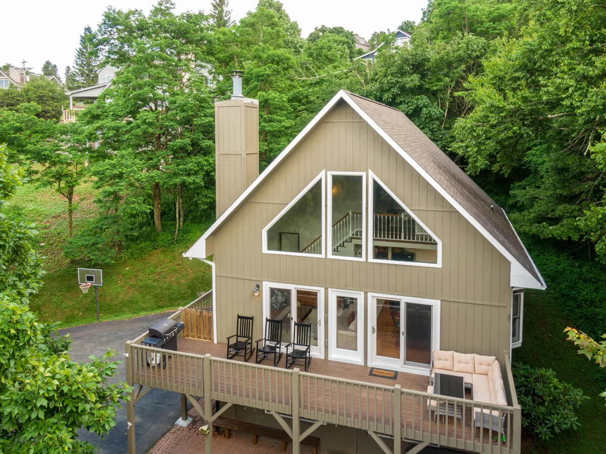 A view of the chalet’s impressive exterior, showcasing large triangular windows, an outdoor seating area on the deck, the deck railing, and a basketball hoop. The lush greenery and ample outdoor space invite recreation and gatherings.