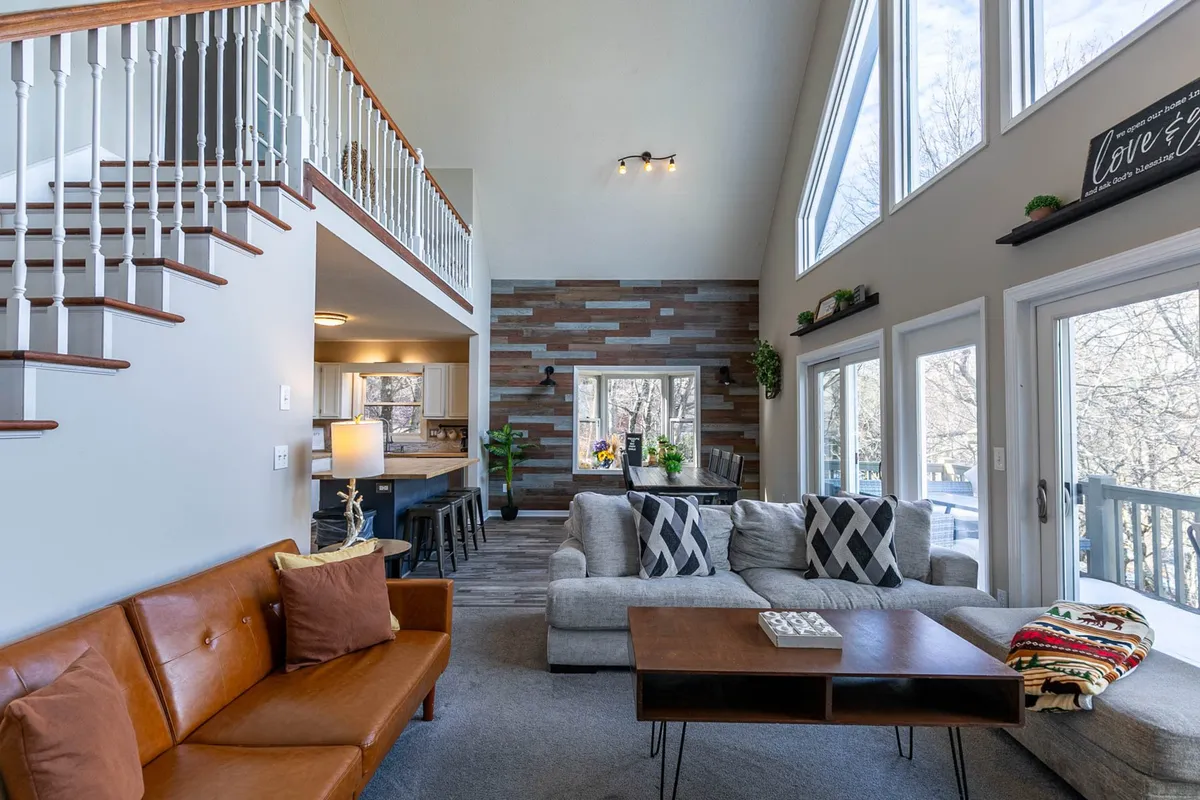 An inviting living area with sunlight pouring in through large windows, cozy seating, and a decorative wooden wall. The windows frame views of nature, making it easy to imagine dogs lounging in the sunlight or guests enjoying the natural surroundings from indoors.