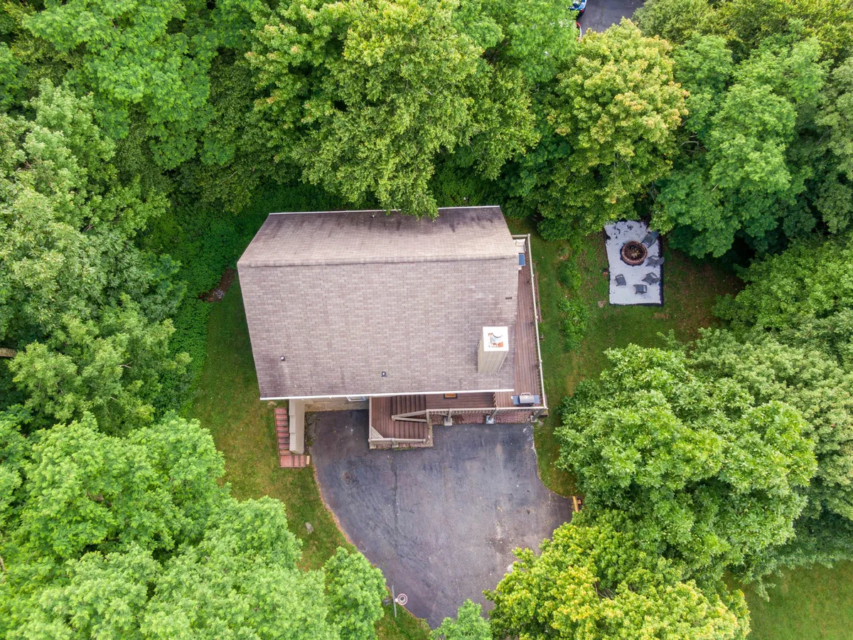 A wide exterior view of the chalet nestled among tall trees, with a focus on the gently sloping yard and a fire pit area surrounded by seating. The driveway is visible, and the natural setting creates a sense of seclusion and connection to nature.