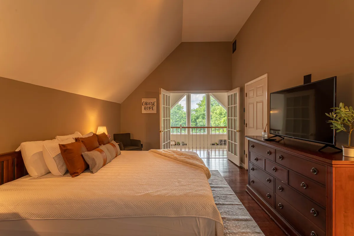 A tranquil bedroom featuring a bed with decorative pillows, large windows offering scenic views, a dresser with a TV, and open balcony doors. The space is softly lit and inviting, perfect for relaxation and quiet retreats.